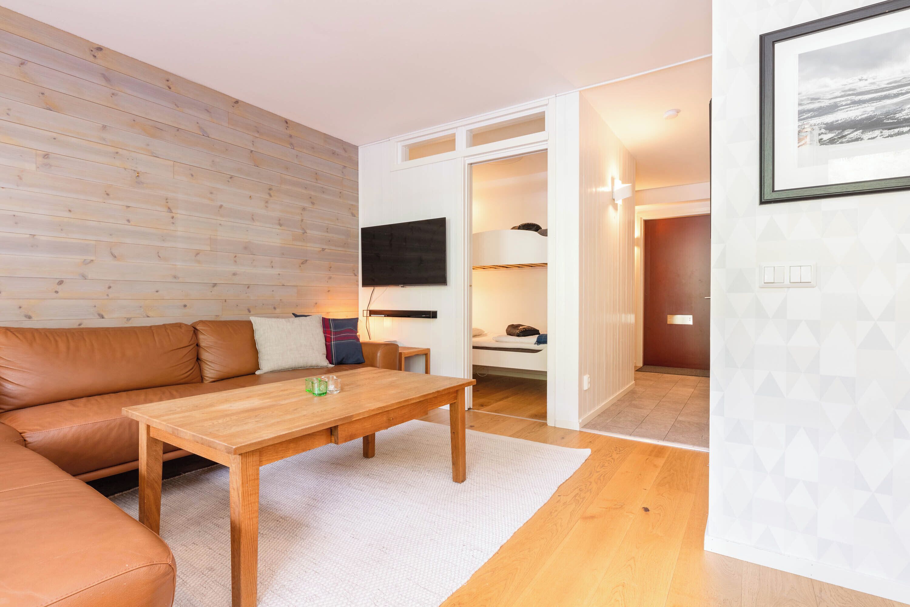Una sala de estar acogedora con piso de madera, un sofá de cuero marrón, una mesa de centro de madera y una pared blanca con un diseño geométrico.