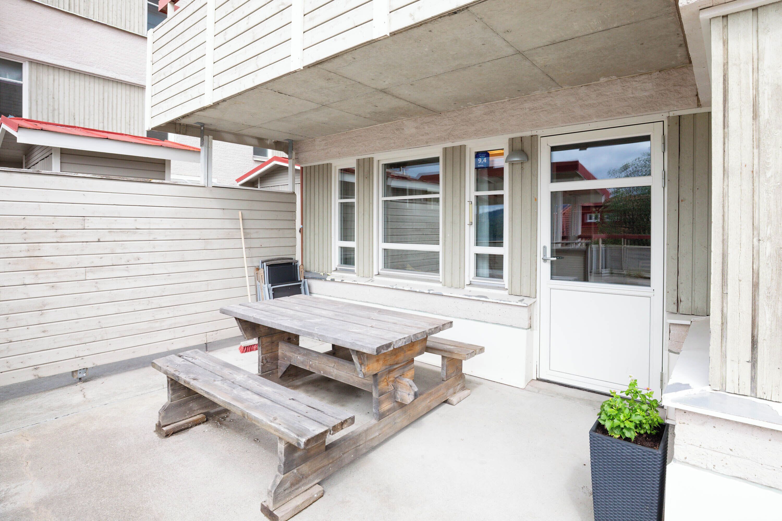 A wooden picnic table and benches outside a building with a white exterior and large windows.