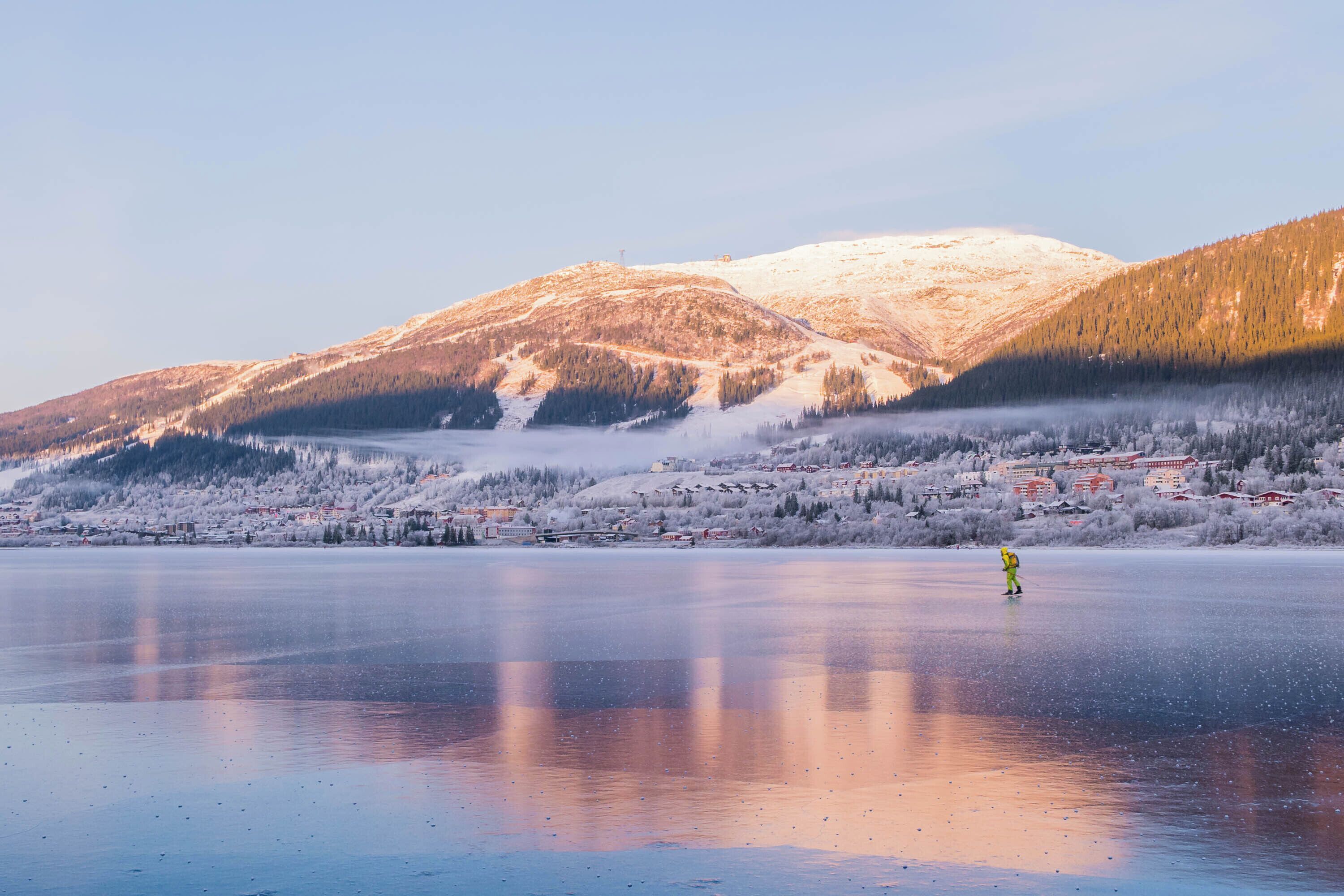 Un lago congelado con una persona vestida de amarillo de pie sobre el hielo, rodeada de montañas cubiertas de nieve y un pueblo al fondo.