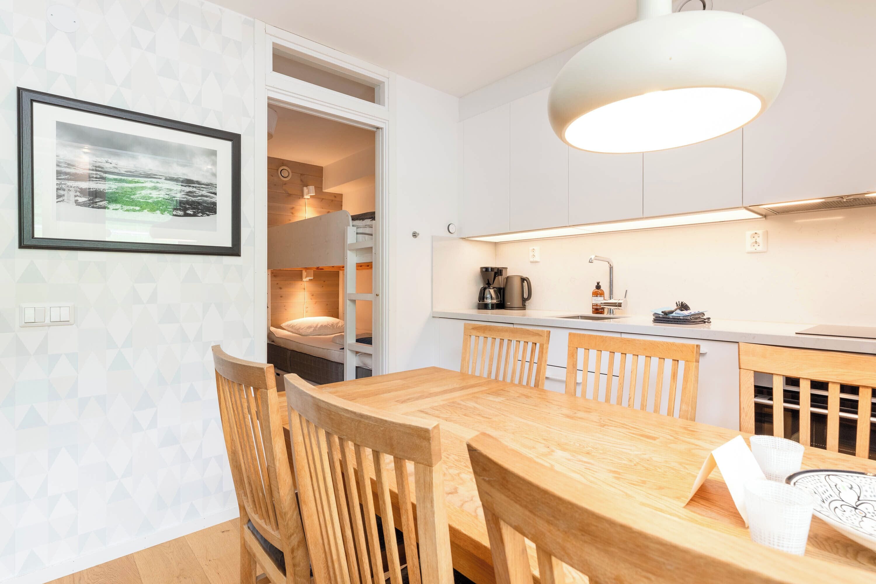 Una cocina moderna con armarios blancos, una mesa de comedor de madera y sillas. Hay un cuadro en la pared, una cafetera y un hervidor en la encimera.