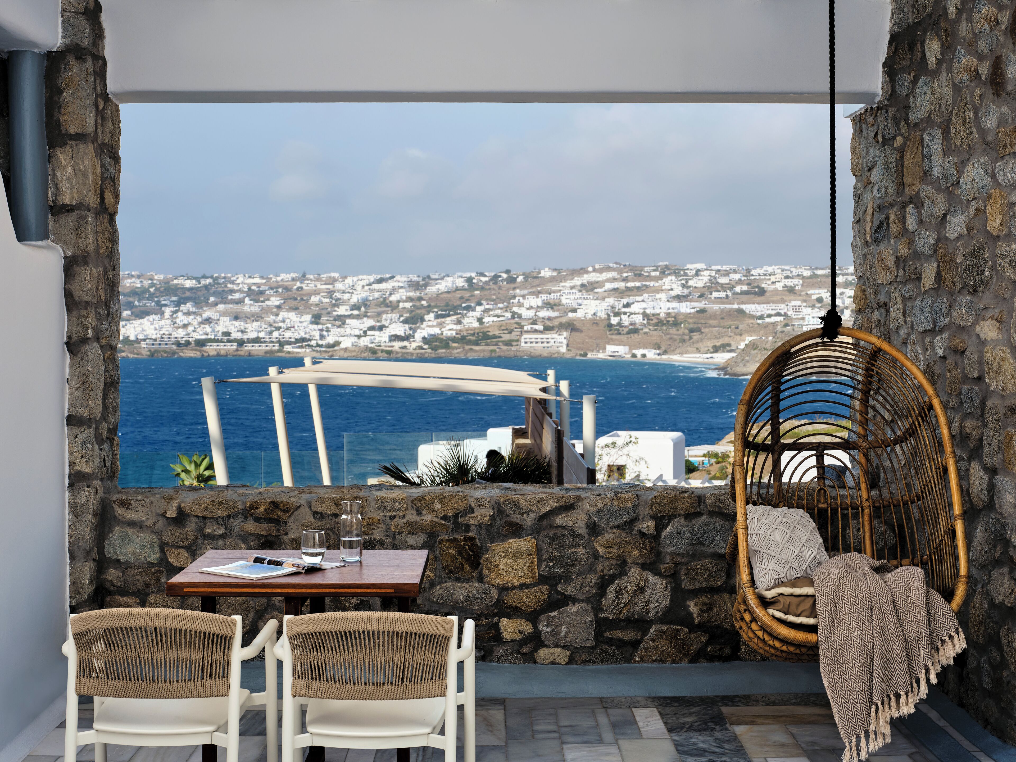 A terrace with a stone wall, a hanging chair, a table with chairs, and a view of the sea and white buildings.