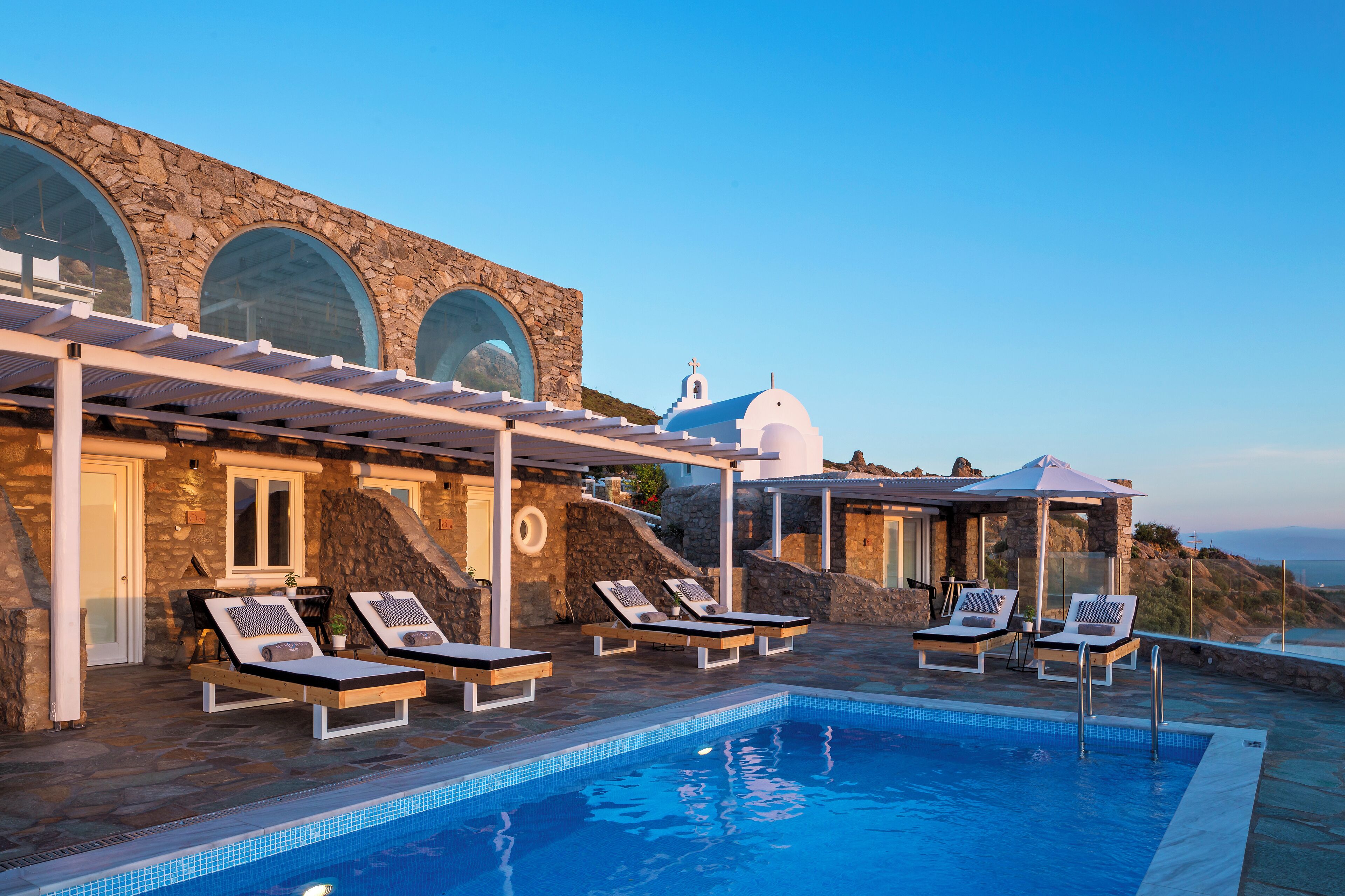 A poolside area with lounge chairs, a stone building with arched windows, and a view of the sea.