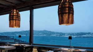 An outdoor dining area with tables and chairs, overlooking a body of water and a mountainous landscape. - Mykonos No5 (Mykonos)