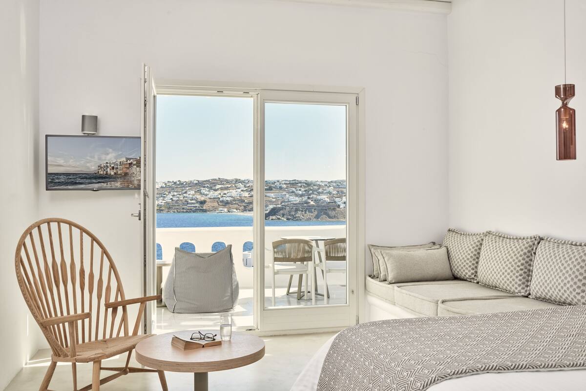 Ein Zimmer mit einem Holzstuhl, einem runden Tisch mit Buch und Gläsern, einer Couch mit gemusterten Kissen und Blick auf eine Küstenlandschaft durch eine Glastür.