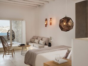 A modern bedroom with a bed, a seating area, a table, and a view of the sea.