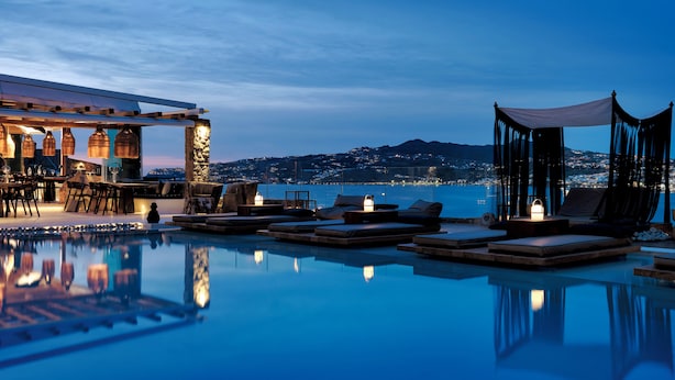 A poolside area with lounge chairs and a bar, overlooking a body of water and a distant cityscape. - Mykonos No5 (Mykonos)