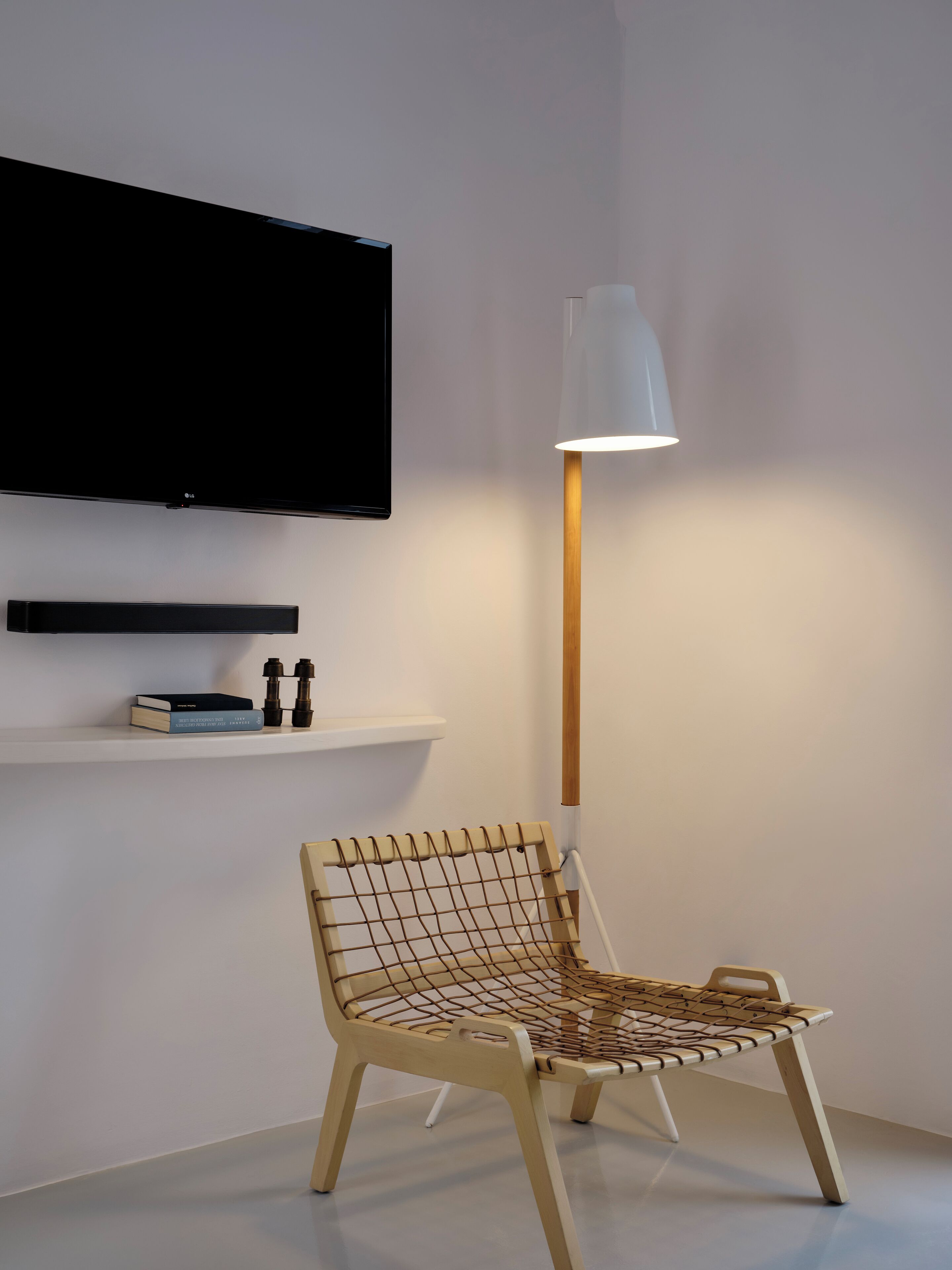 Une chambre moderne avec un téléviseur fixé au mur, une lampe sur pied, une chaise avec un siège en osier et une étagère contenant des livres et des jumelles.