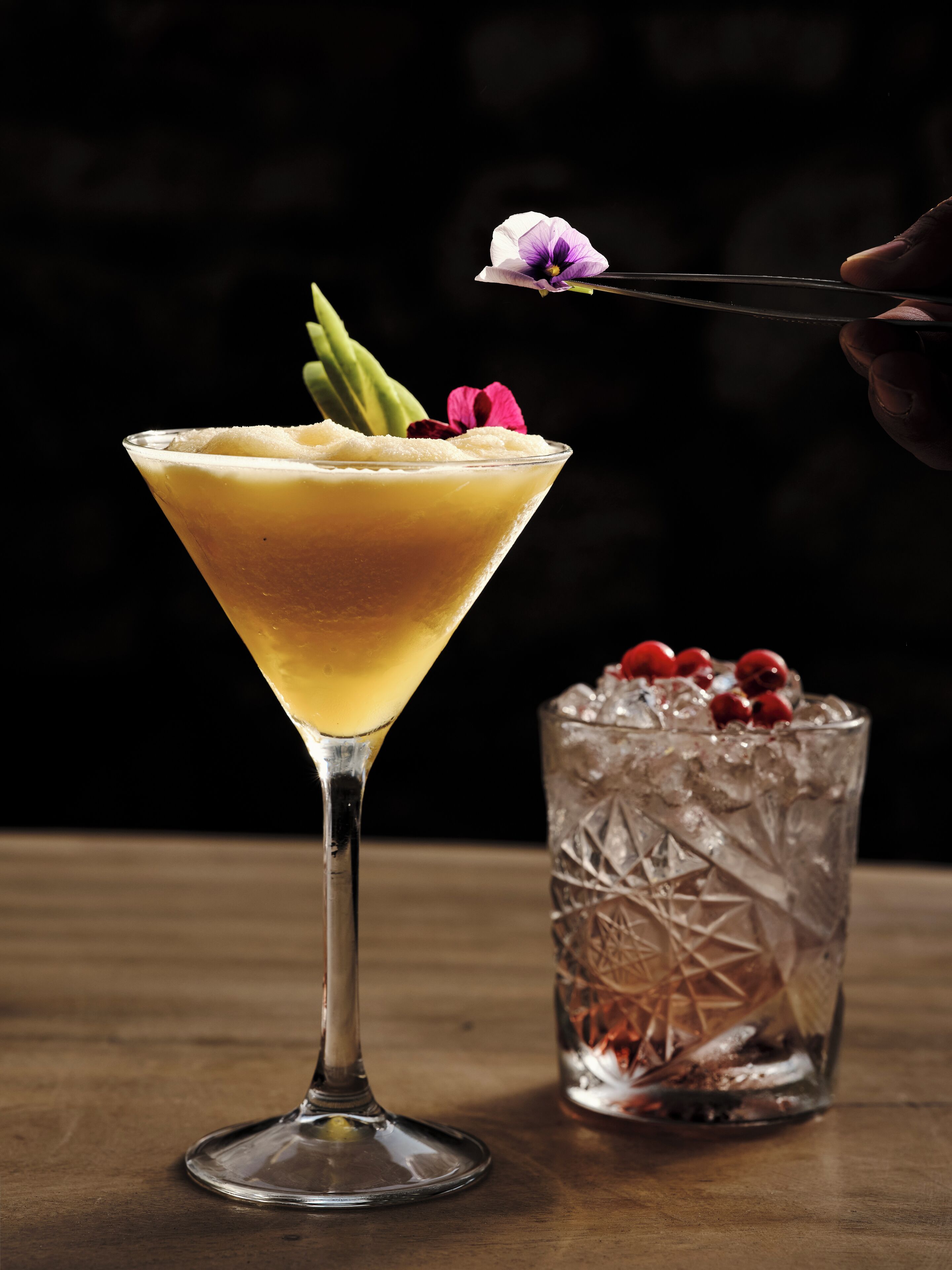 A martini glass with a yellowish drink, garnished with a flower and a sprig of greenery, on a wooden surface.
