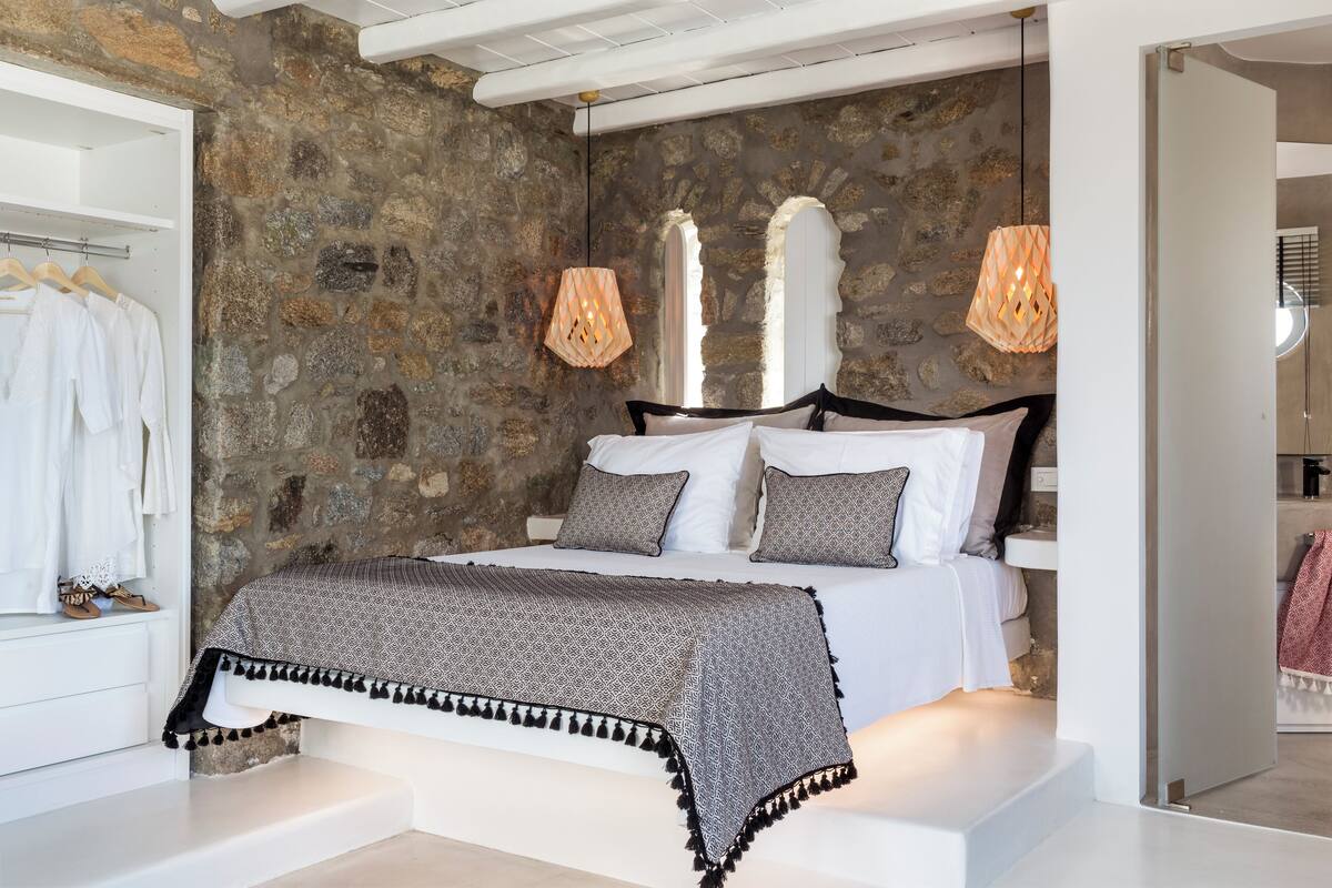 Ein Schlafzimmer mit einer Steinwand, einem Bett mit gemusterten Betttextilien und zwei Pendelleuchten.