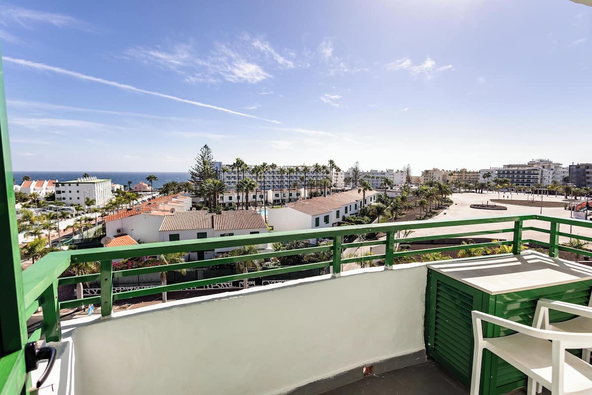 Ein Balkon mit grüner Geländer, der auf eine Küstenlandschaft mit Gebäuden und Palmen blickt.