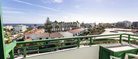 A balcony with a green railing overlooking a coastal area with buildings and palm trees.