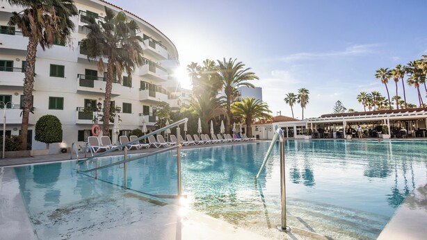 A pool area with lounge chairs and palm trees, surrounded by a multi-story building with balconies. - Servatur Playa Bonita (San Bartolomé de Tirajana)