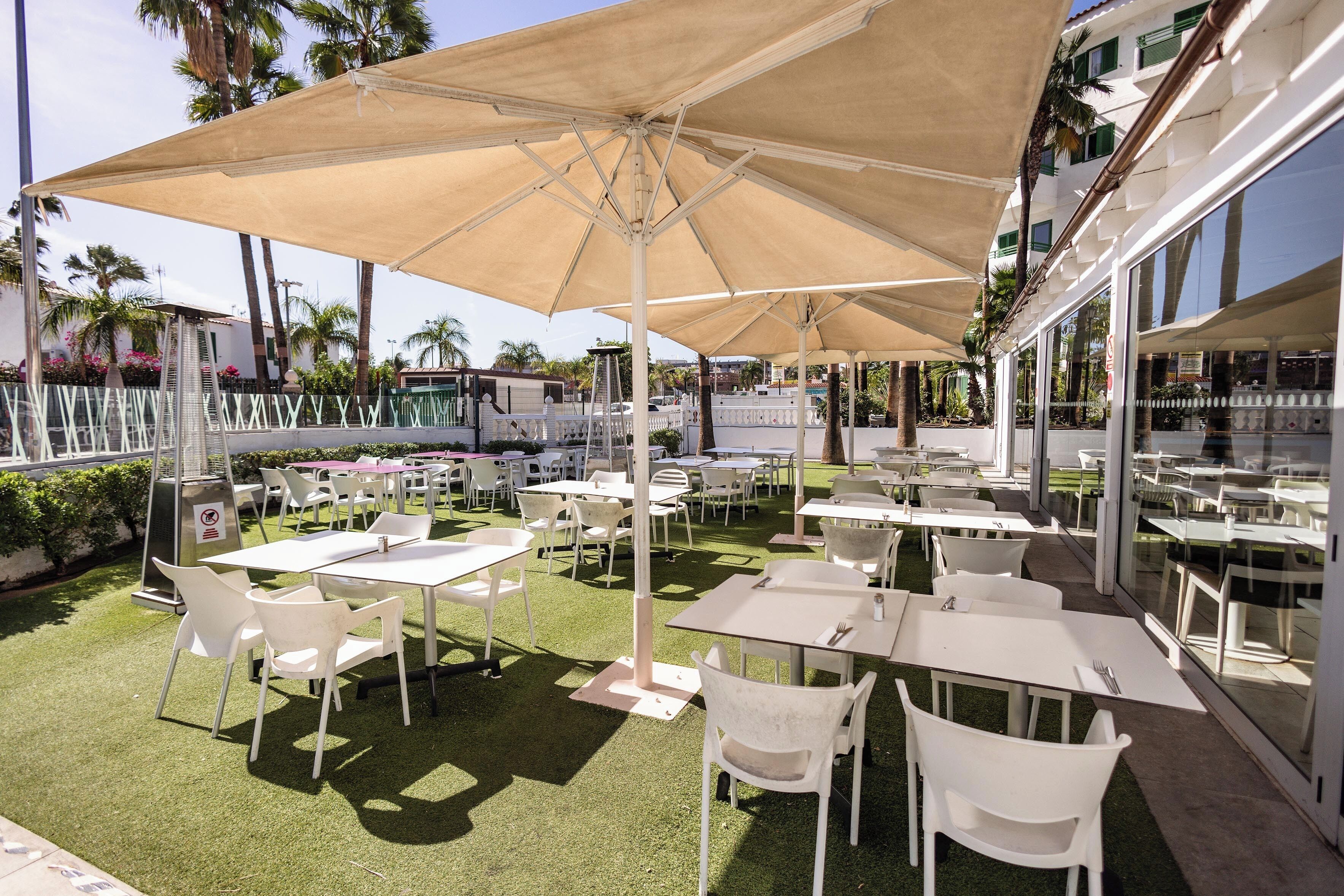 An outdoor patio with white tables and chairs, shaded by large umbrellas, and surrounded by palm trees.