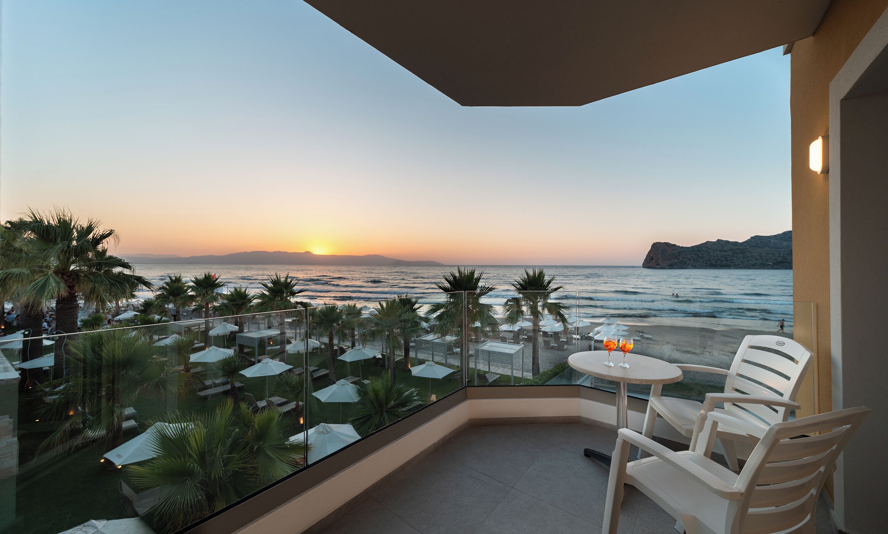 A balcony with a view of the sea at sunset, outdoor seating, and umbrellas.