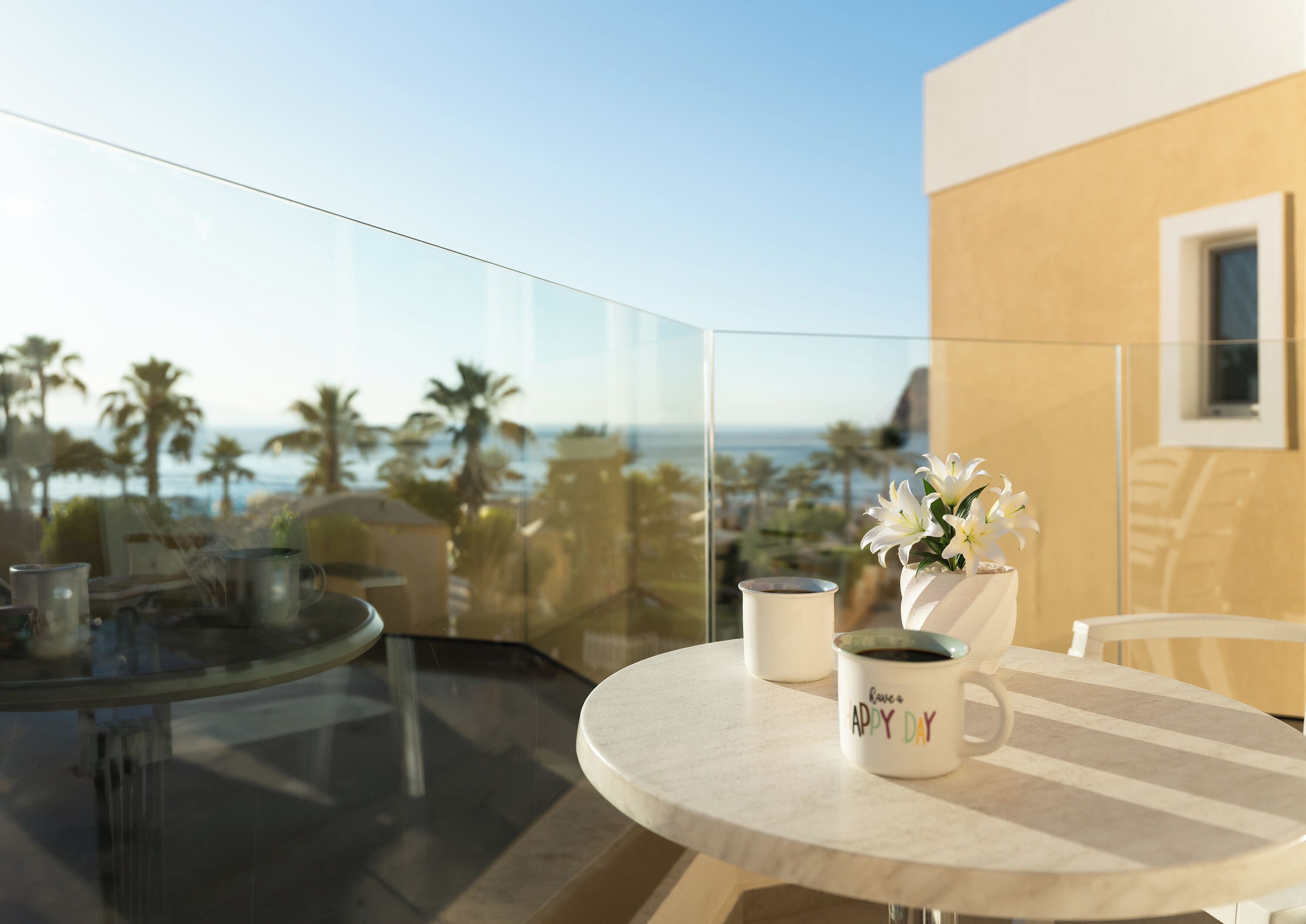 A balcony with a glass railing, a round table with two white mugs, and a vase with white flowers.