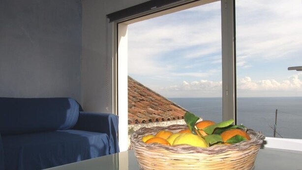 Une pièce avec une table en verre, un panier en osier rempli de fruits, un canapé bleu et une vue sur la mer par une grande fenêtre.