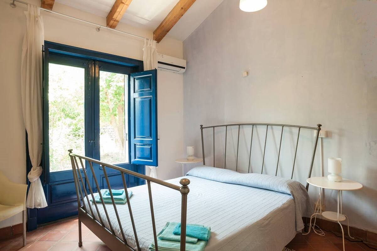 A bedroom with a bed, a window with curtains, a bedside table, and a small table with a lamp.