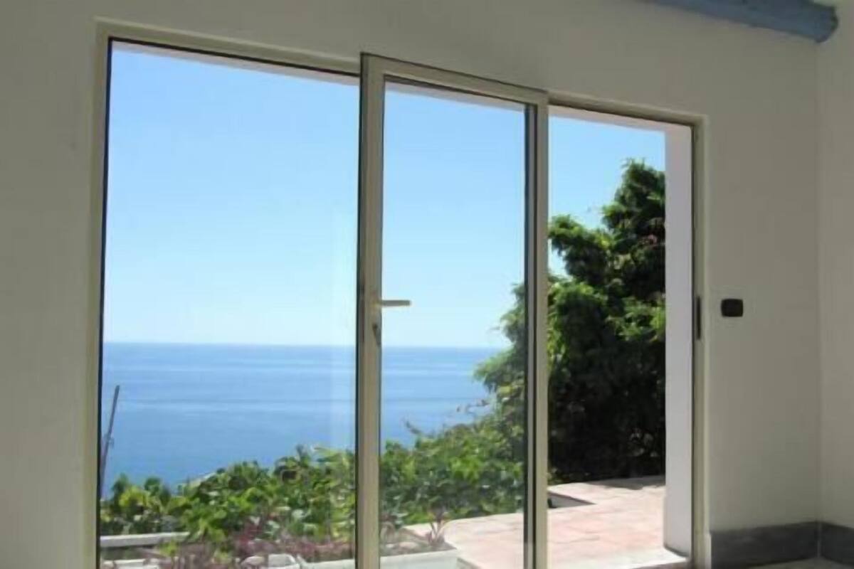 A room with a sliding glass door leading to a balcony with a view of the sea and greenery.