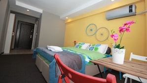 Quarto de hotel com cama, mesa e cadeira. Há uma porta e um aparelho de ar condicionado na parede.