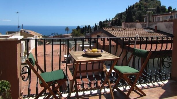 A balcony with a table and chairs, overlooking a coastal view with buildings and trees. - Come In Sicily - Al Mercato (Taormina)