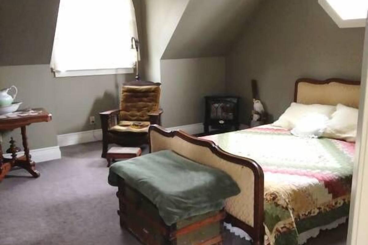 Une chambre avec un plafond en pente, un lit recouvert d’une courtepointe, une chaise, une petite table et une fenêtre.