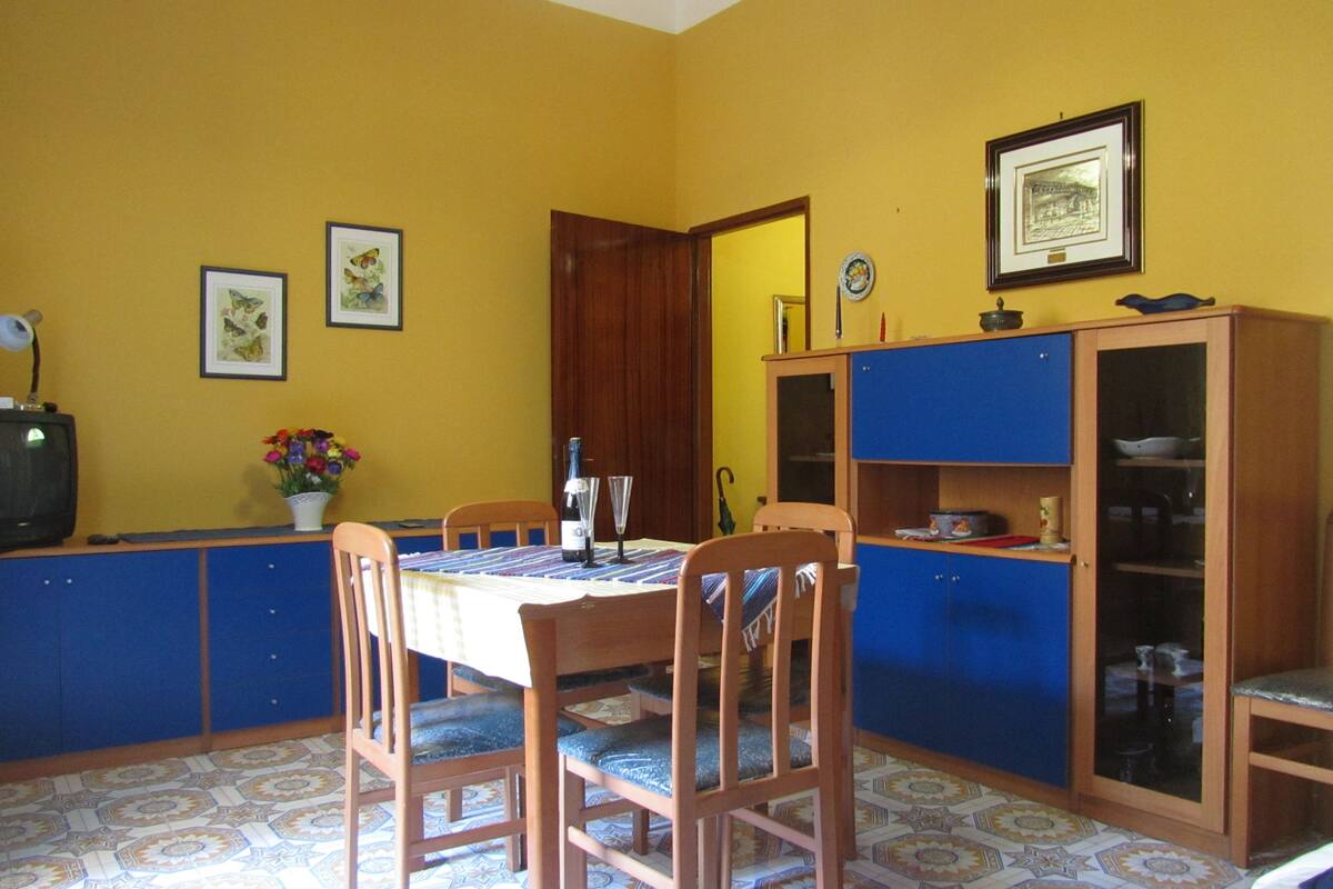 A dining area with a table set for two, wooden chairs, a cabinet with glass doors, and a television on a stand.