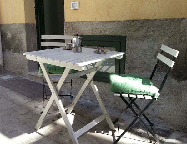 Une petite table d’extérieur avec un pot et des tasses en métal, une chaise rembourrée verte et un banc vert adossé à un mur à texture.
