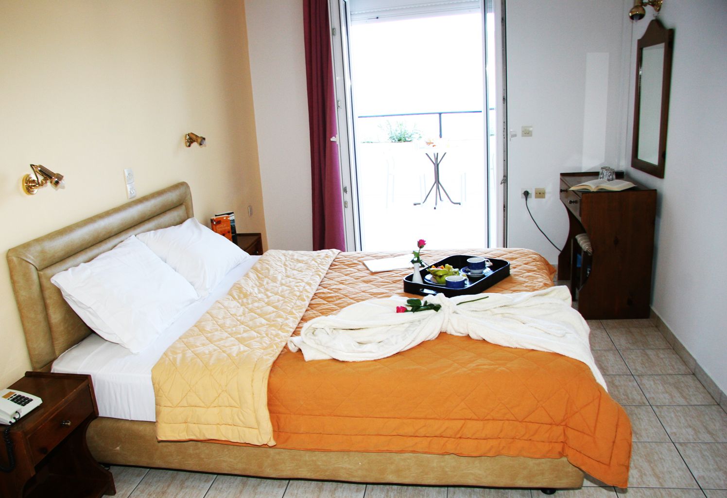 Habitación de hotel con cama, mesita de noche con teléfono, ventana con cortinas, una pequeña mesa con una bandeja de alimentos para el desayuno y un espejo en la pared.