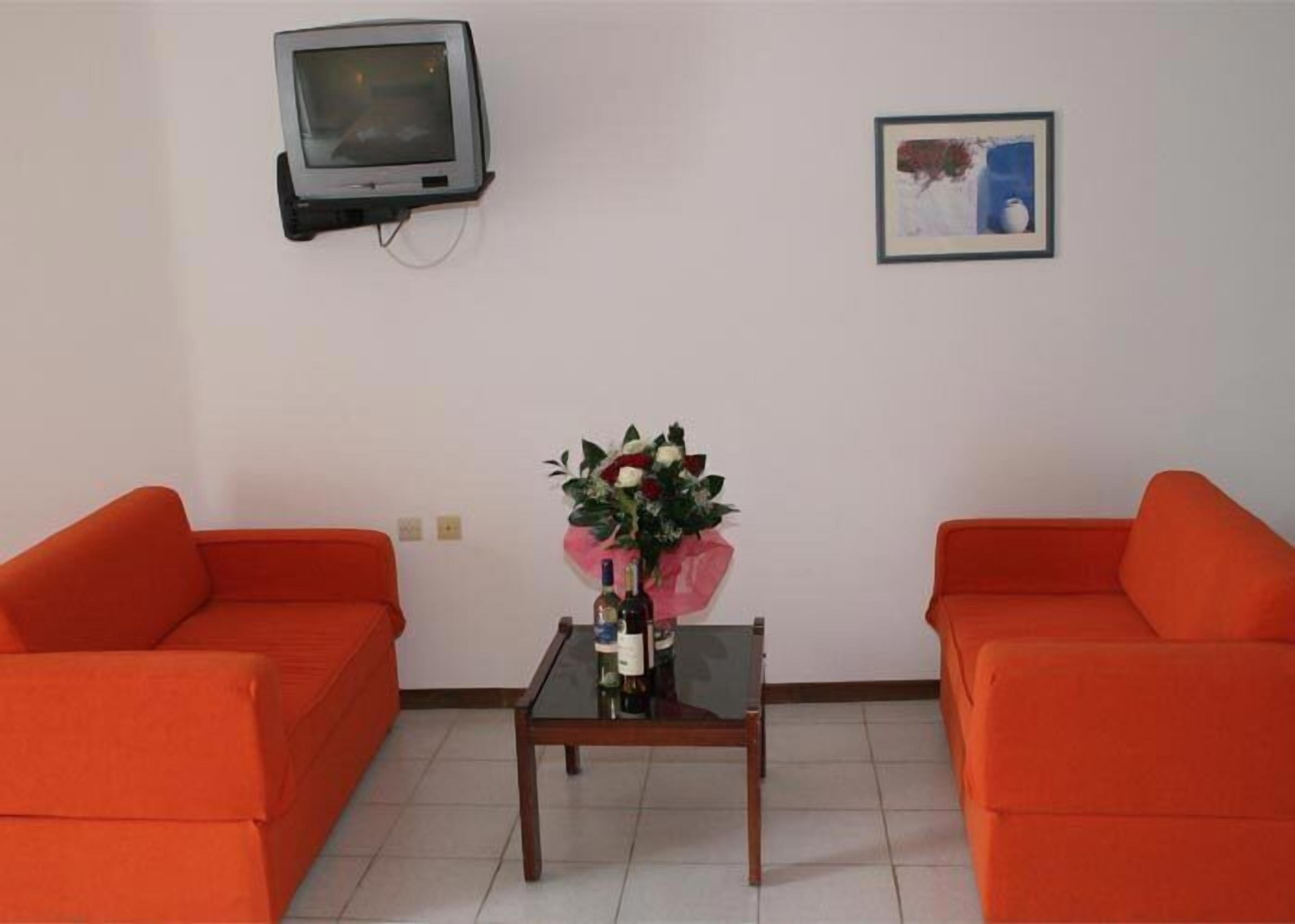 Una habitación con dos sofás naranja, una mesita con un florero, un televisor montado en la pared y un cuadro enmarcado.