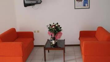 Una habitación con dos sofás naranja, una mesita con un florero, un televisor montado en la pared y un cuadro enmarcado.