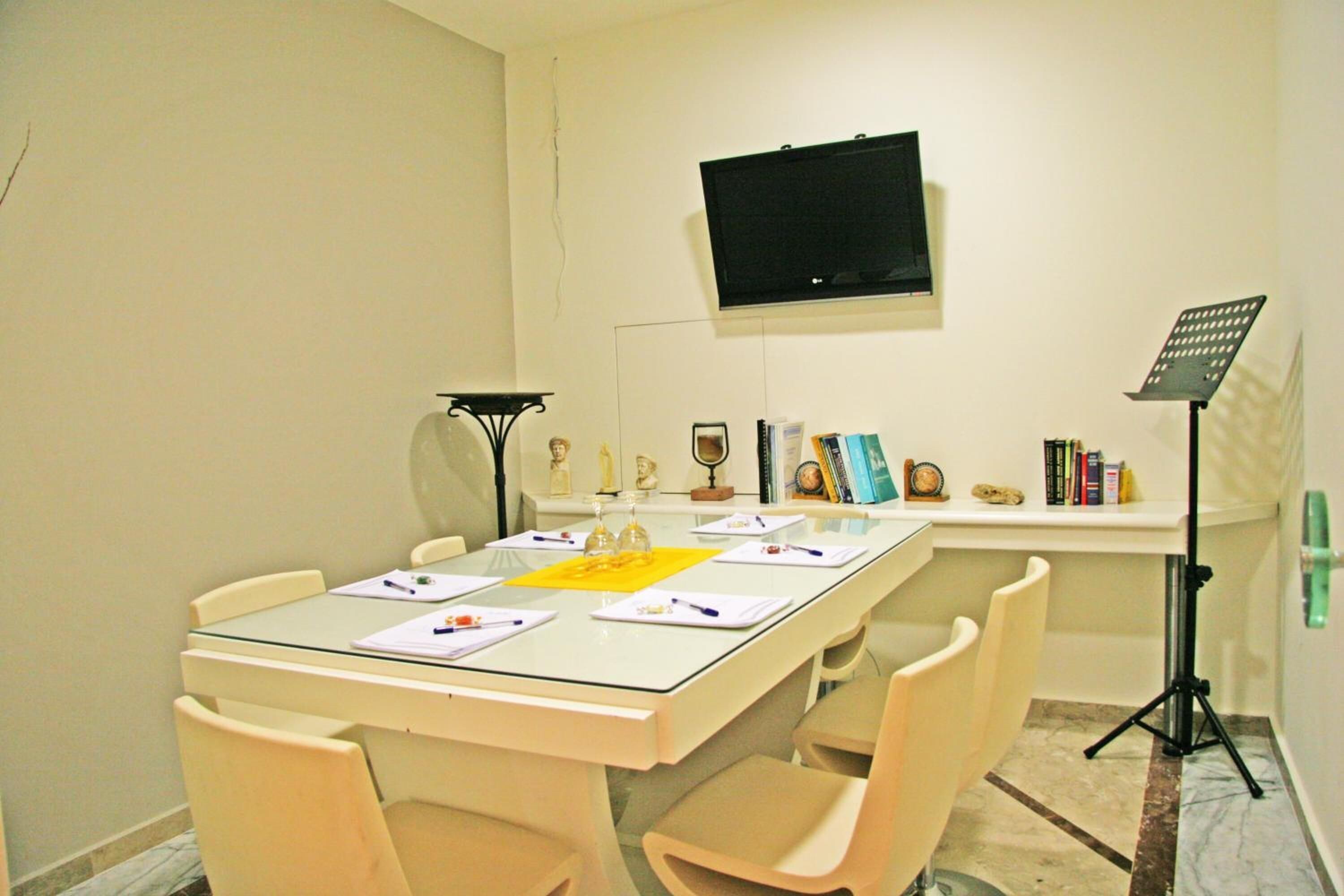 A conference room with a glass table, beige chairs, a mounted TV, a music stand, and a tripod.