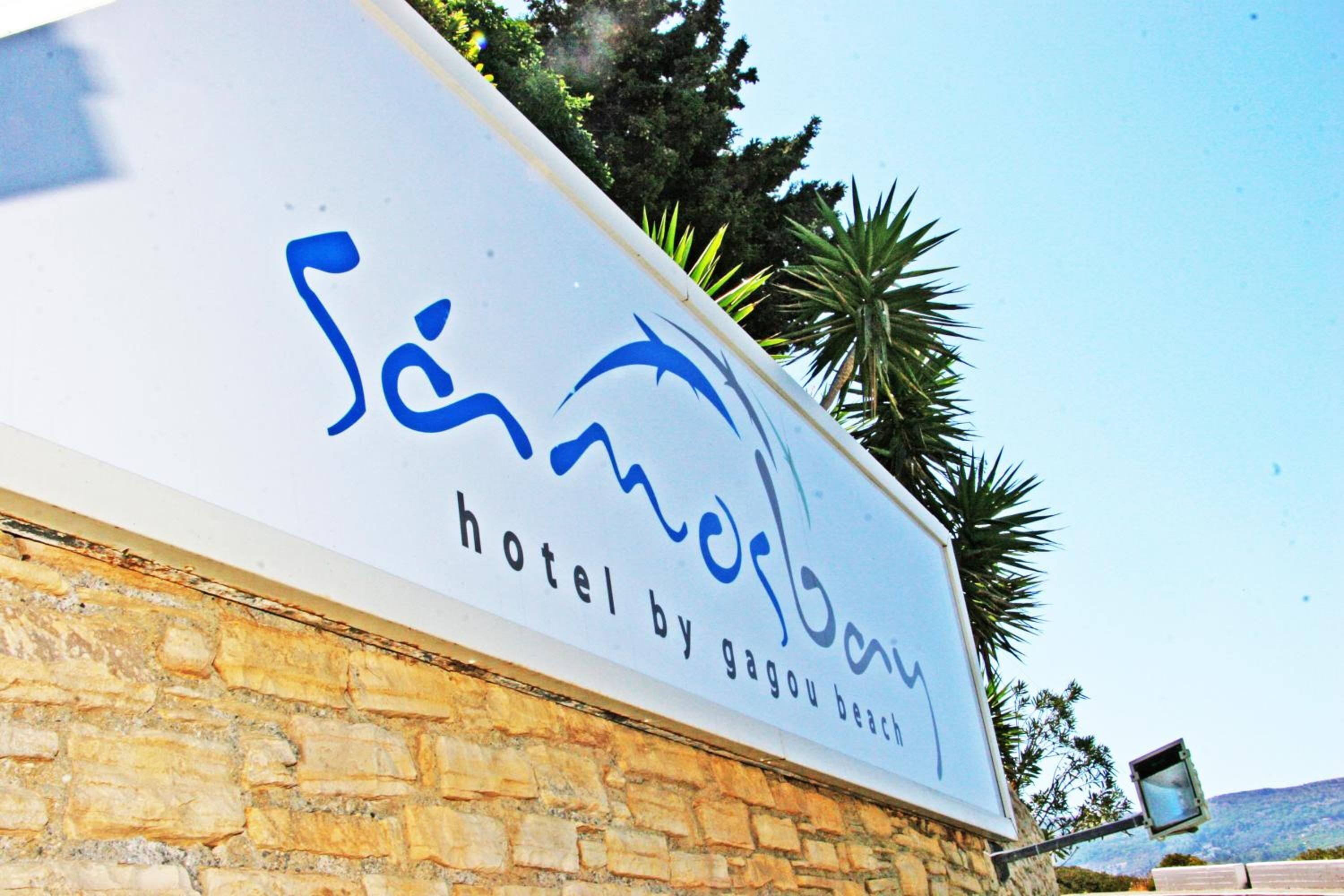 A sign for 'Samanalao Hotel by gagou beach' with a stone wall and palm trees in the background.