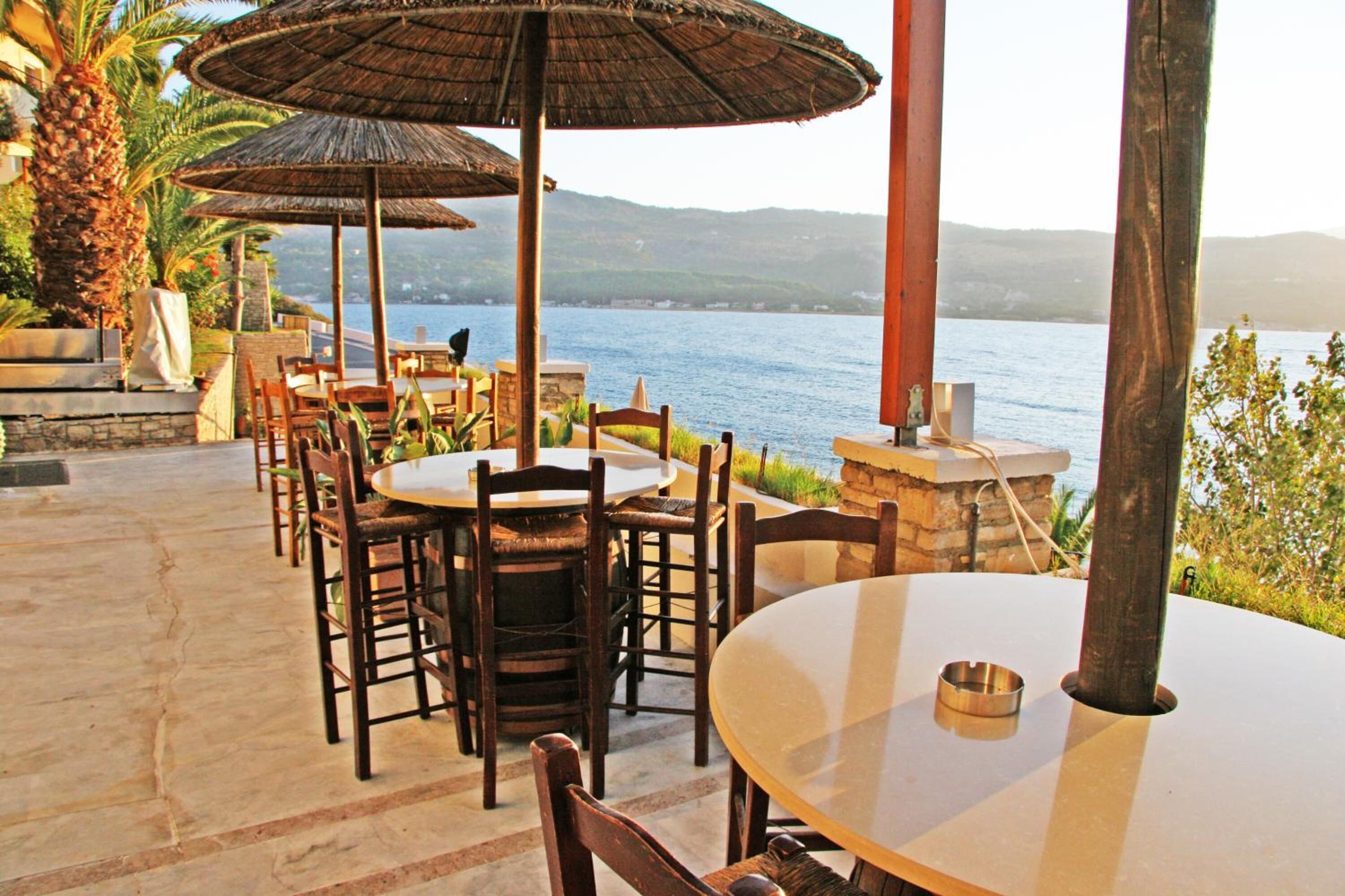 Un espace de restauration en plein air, avec des tables et des chaises rondes, entouré de murs en pierre et surplombé de parasols en chaume, donnant sur une étendue d’eau.
