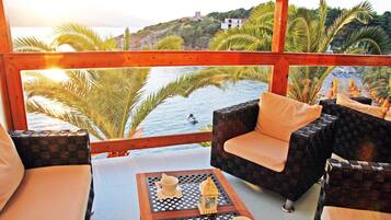 Ein Balkon mit Korbmöbeln, einem Tisch mit Tasse und kleiner Laterne sowie Blick auf das Meer und einen Hügel mit Gebäuden.