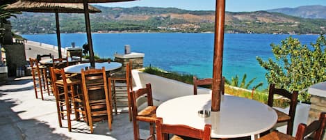 An outdoor seating area with round tables and wooden chairs, overlooking a body of water and hills.