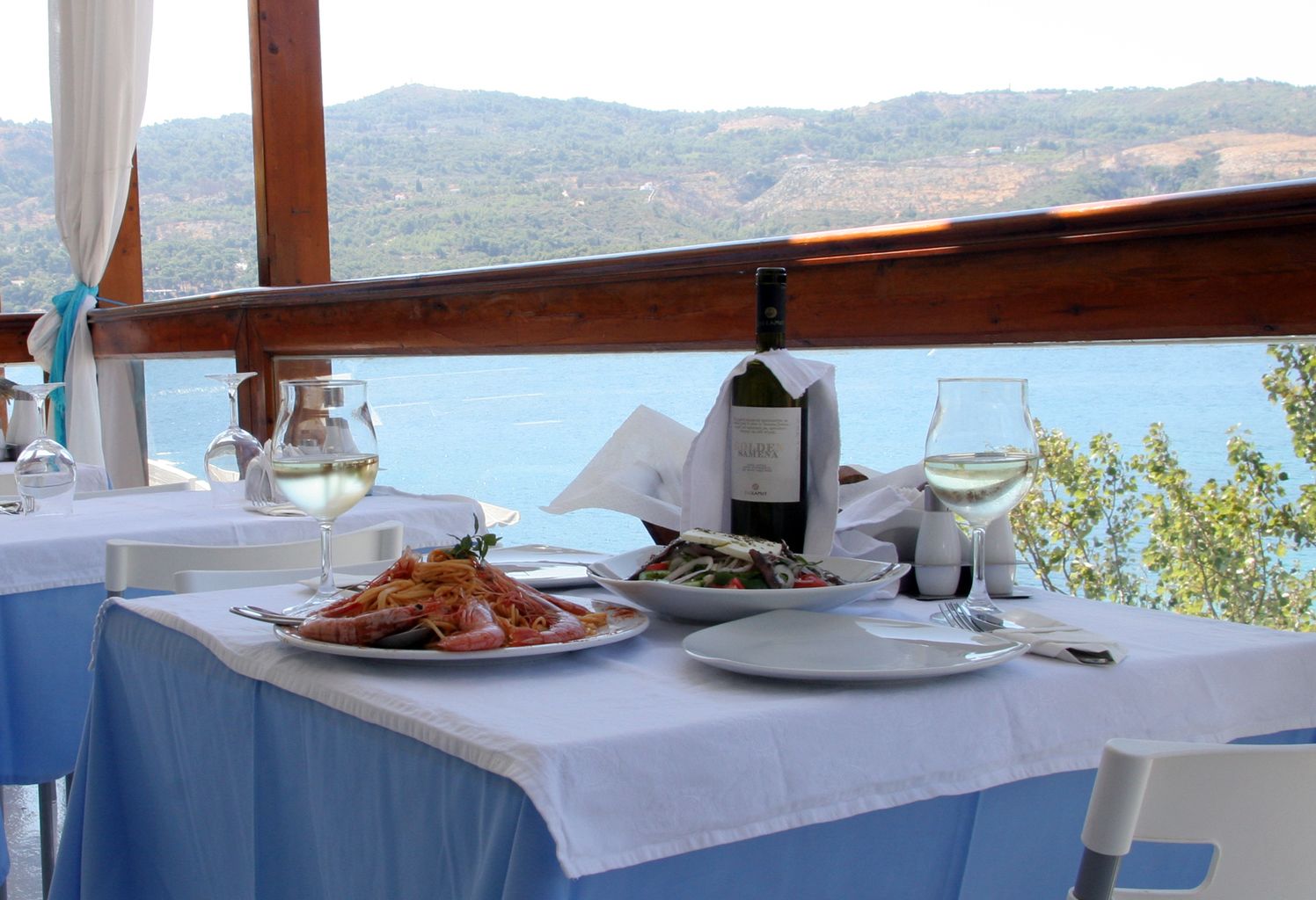 Une table à manger dressée avec des plats de fruits de mer, des verres à vin et une bouteille de vin, offrant une vue panoramique sur des collines et l’eau.