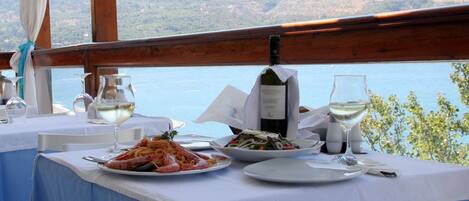 Une table à manger dressée avec des plats de fruits de mer, des verres à vin et une bouteille de vin, offrant une vue panoramique sur des collines et l’eau.