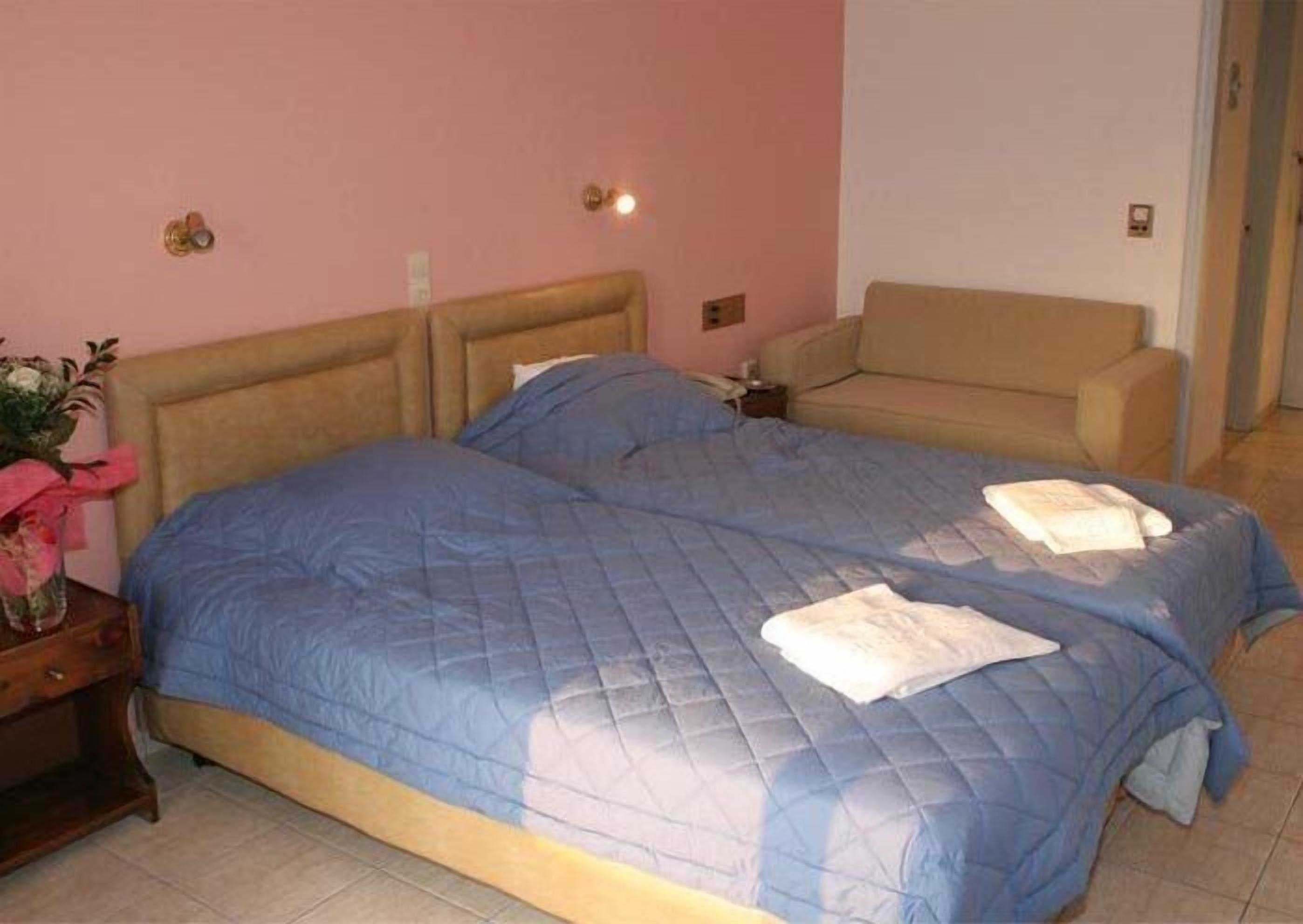 Uma cama de casal com um edredom azul, um sofá bege e uma pequena mesa de cabeceira com um vaso de flores.