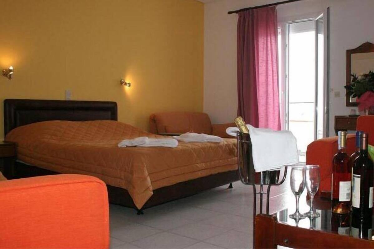 A hotel room with a bed, a chair, a table with wine glasses and bottles, and a window with curtains.