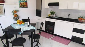 A compact kitchen with white cabinets, a black countertop, and a dining area with a table and chairs. - Come In Sicily - Beachfront Sant'Alessio (Sant'Alessio Siculo)