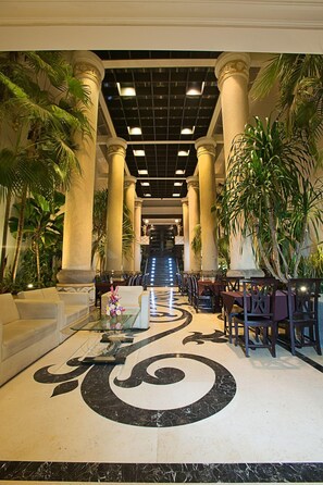 Un vestíbulo con una escalera central, flanqueada por columnas y palmeras, que conduce a un piso superior.