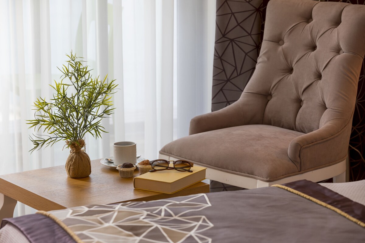 Une pièce comprenant un fauteuil capitonné, une table en bois ornée d’un vase de plantes vertes, un livre, une paire de lunettes et un coussin décoratif.