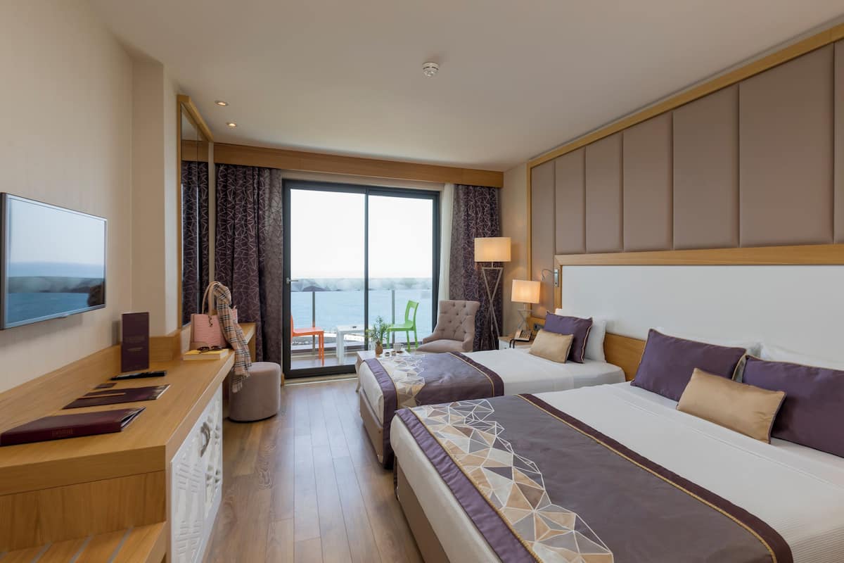A hotel room with two beds, a TV, a desk with a computer, a chair, and a view of the sea.
