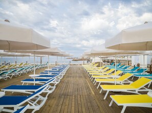 A beachside deck with rows of sun loungers under umbrellas, a wooden walkway, and a view of the sea and distant shoreline. - Mary Hotels Alanya - All inclusive (Alanya)