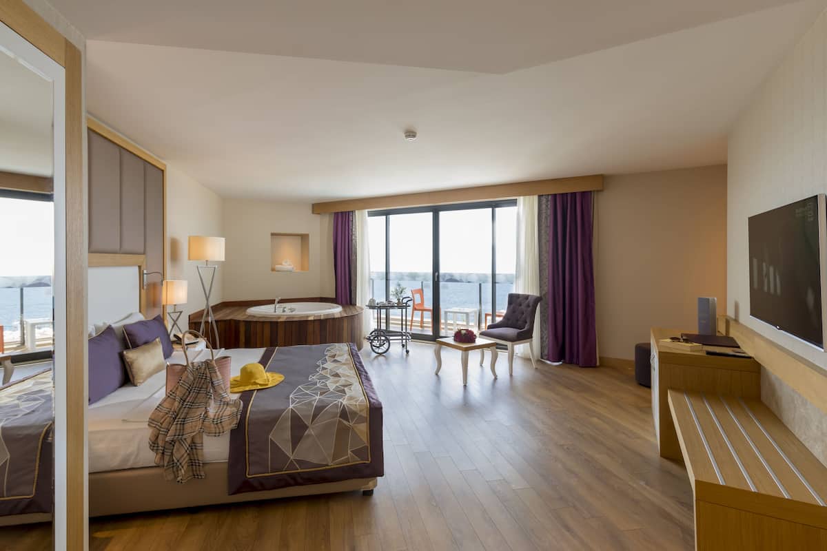 A hotel room with a bed, a desk, a chair, a small table, a sink, and a large window with a view of the sea.