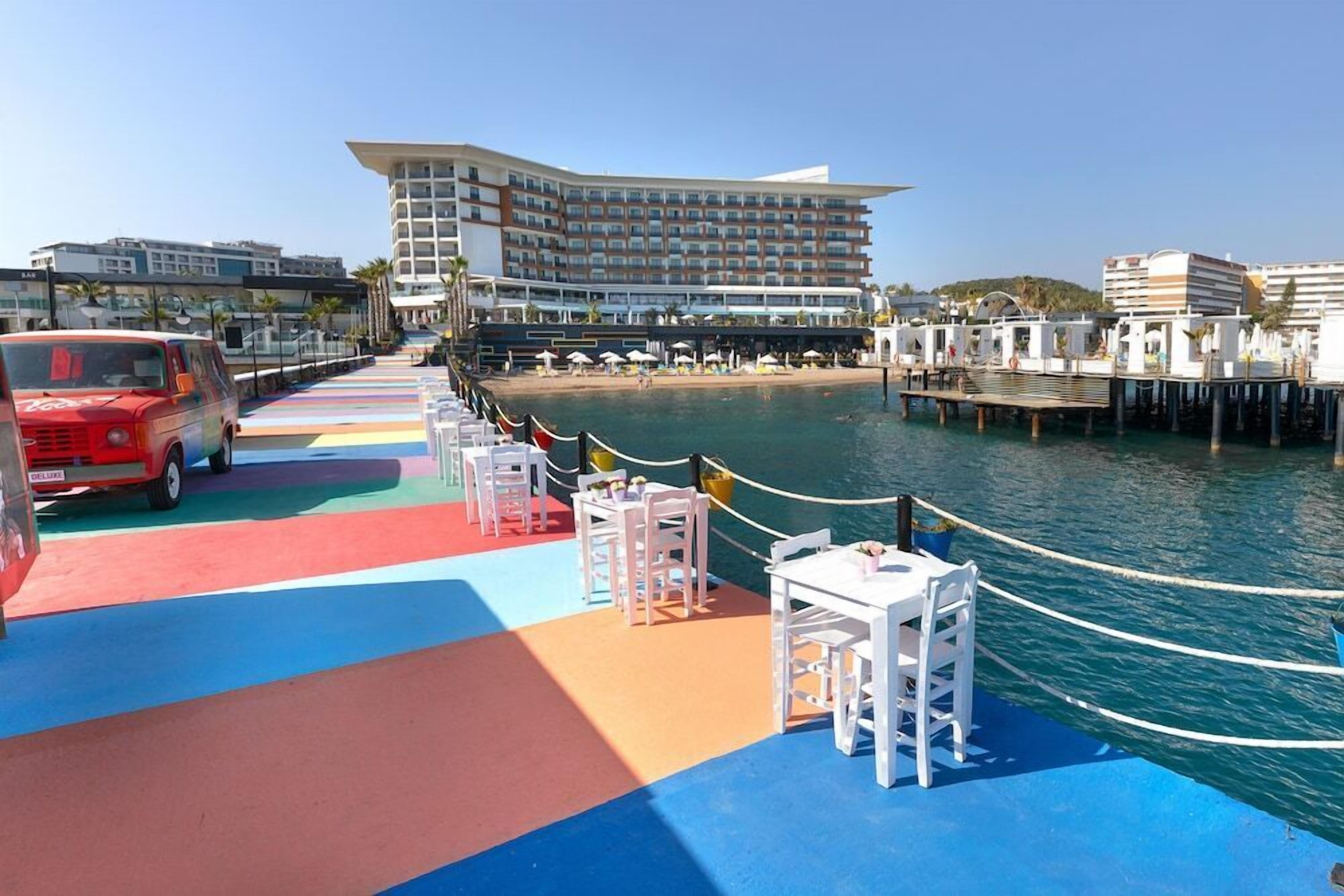 A waterfront promenade with colorful seating areas, a red vehicle, and a large hotel building in the background.