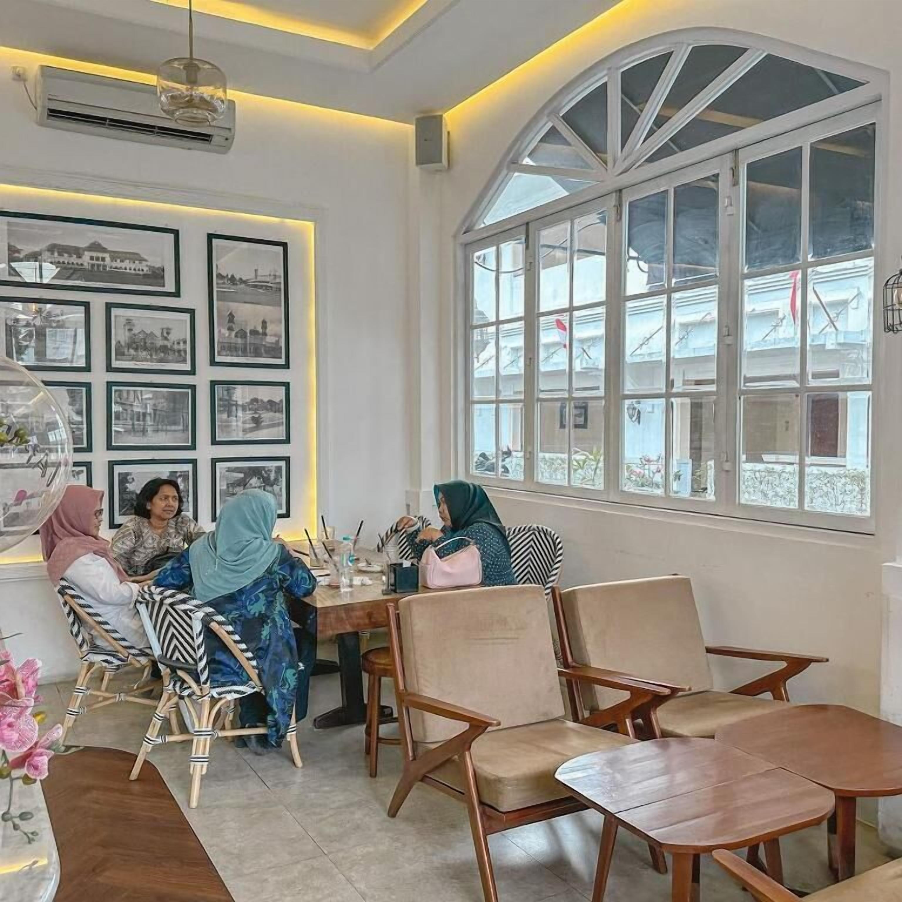 Una habitación con un ventanal arqueado grande, una pared con cuadros enmarcados y un grupo de personas sentadas en una mesa.