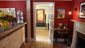 A wooden hallway with a red wall, a yellow vase with flowers, a framed picture, and a table with bottles. - King Hill Inn & Kitchen (New London)