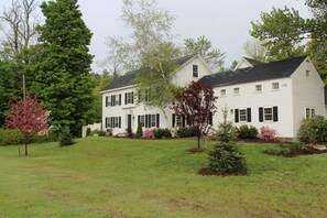 Une maison blanche à deux étages, dotée de volets noirs, entourée d’une pelouse bien entretenue et de divers arbres.