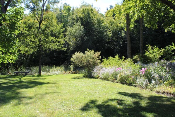 A well-maintained garden with a grassy lawn, various flowering plants, and tall trees. - King Hill Inn & Kitchen (New London)